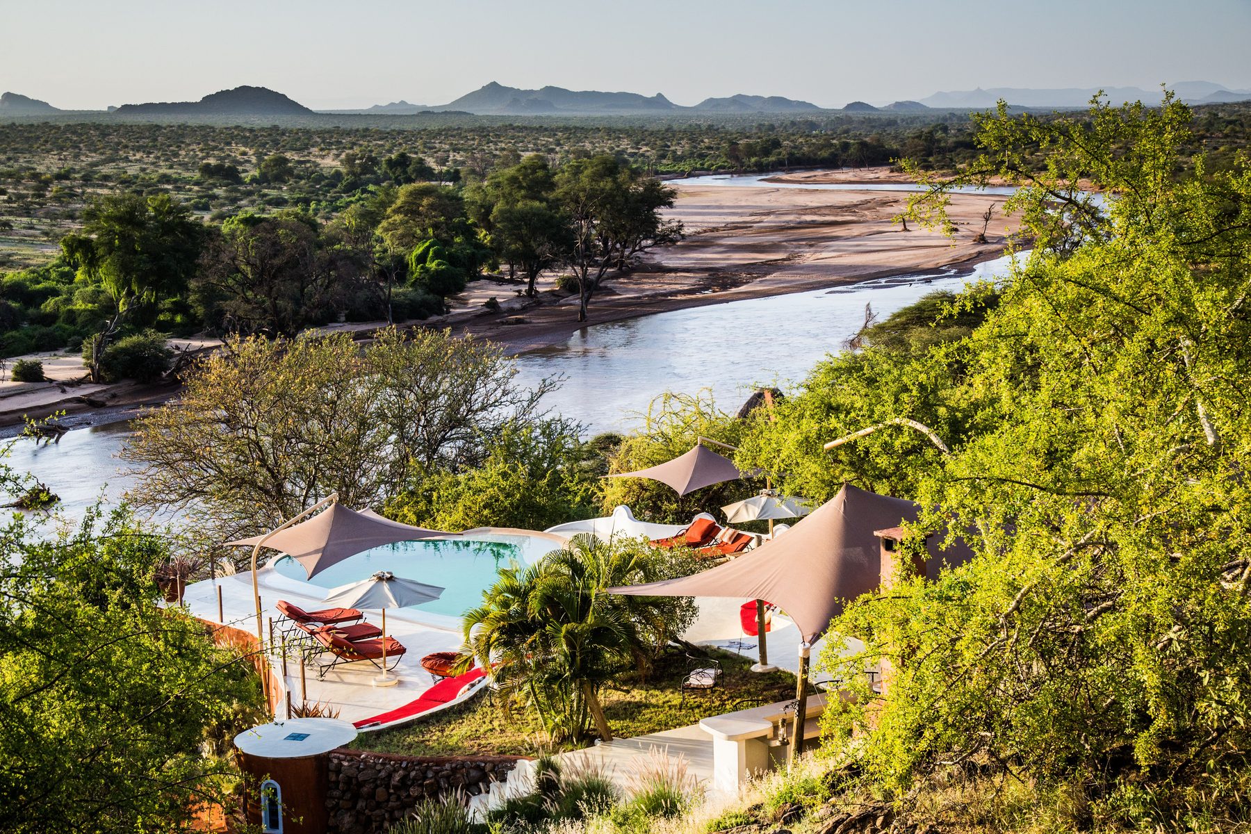 Aerial view of Sasaab’s pool and umbrellas among green trees above a wide sandy river, hills in the distance