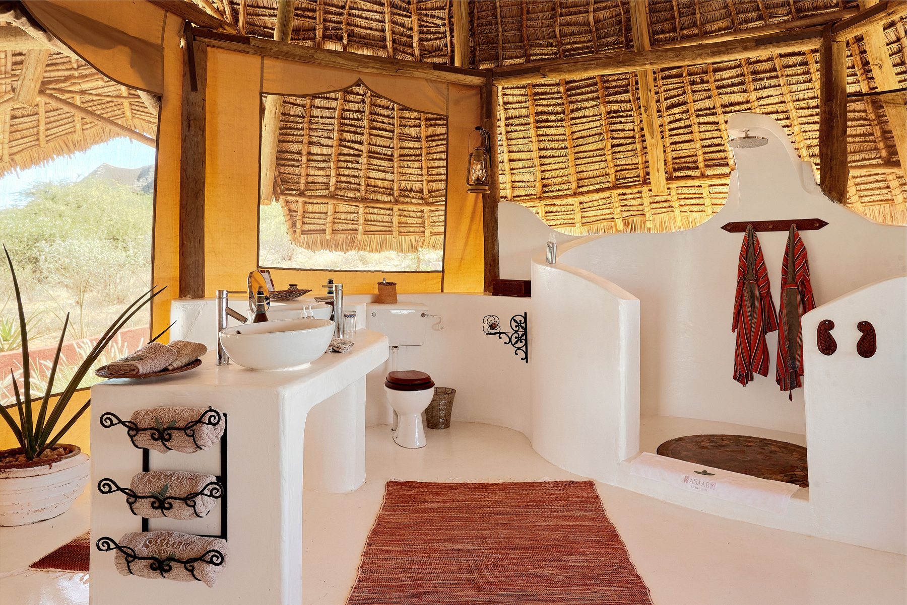 White plastered open bathroom at Sasaab with a basin, red towels and a shower under a woven thatched ceiling