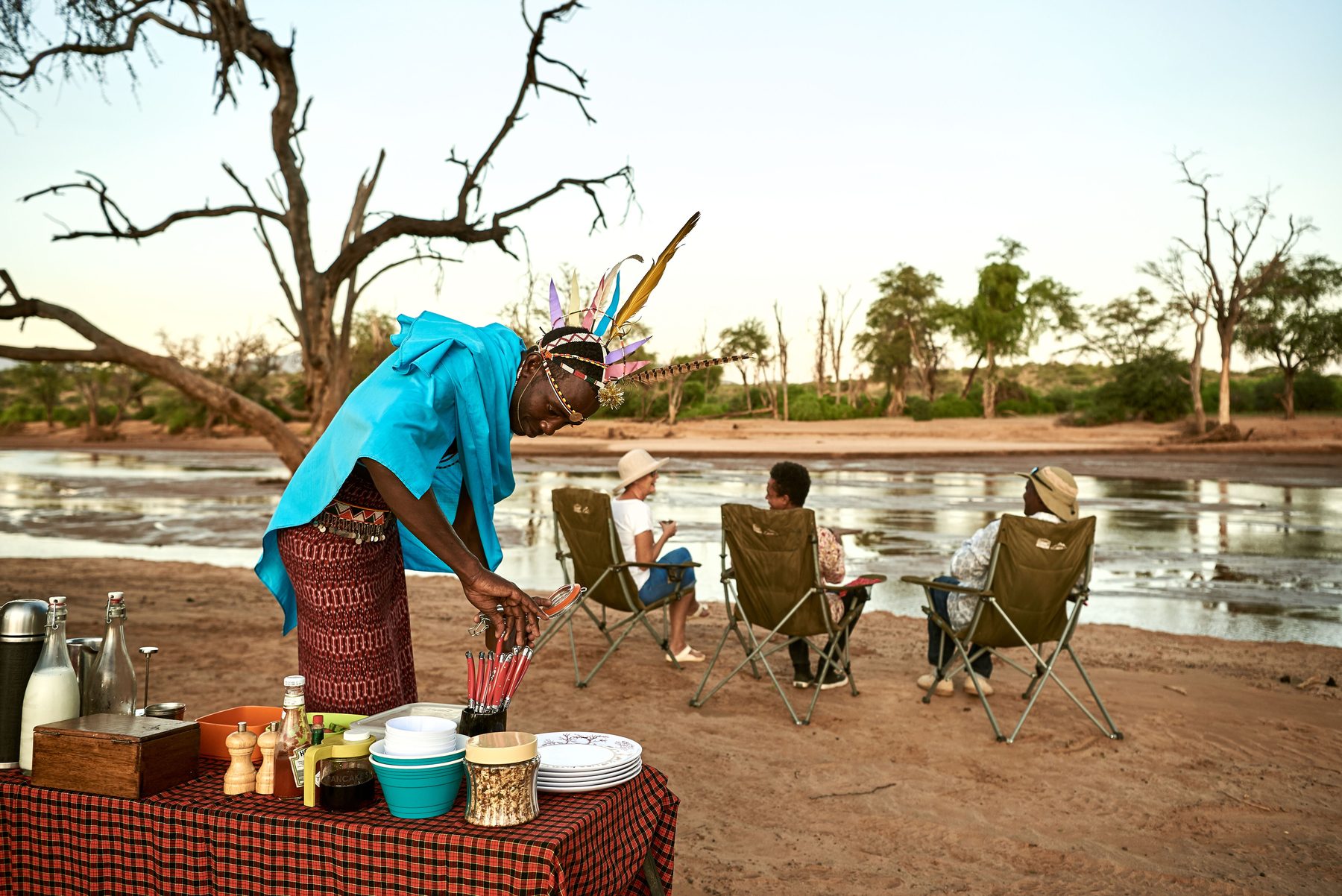 Guide in Samburu dress preparing a meal at a table beside the river as guests relax in camp chairs