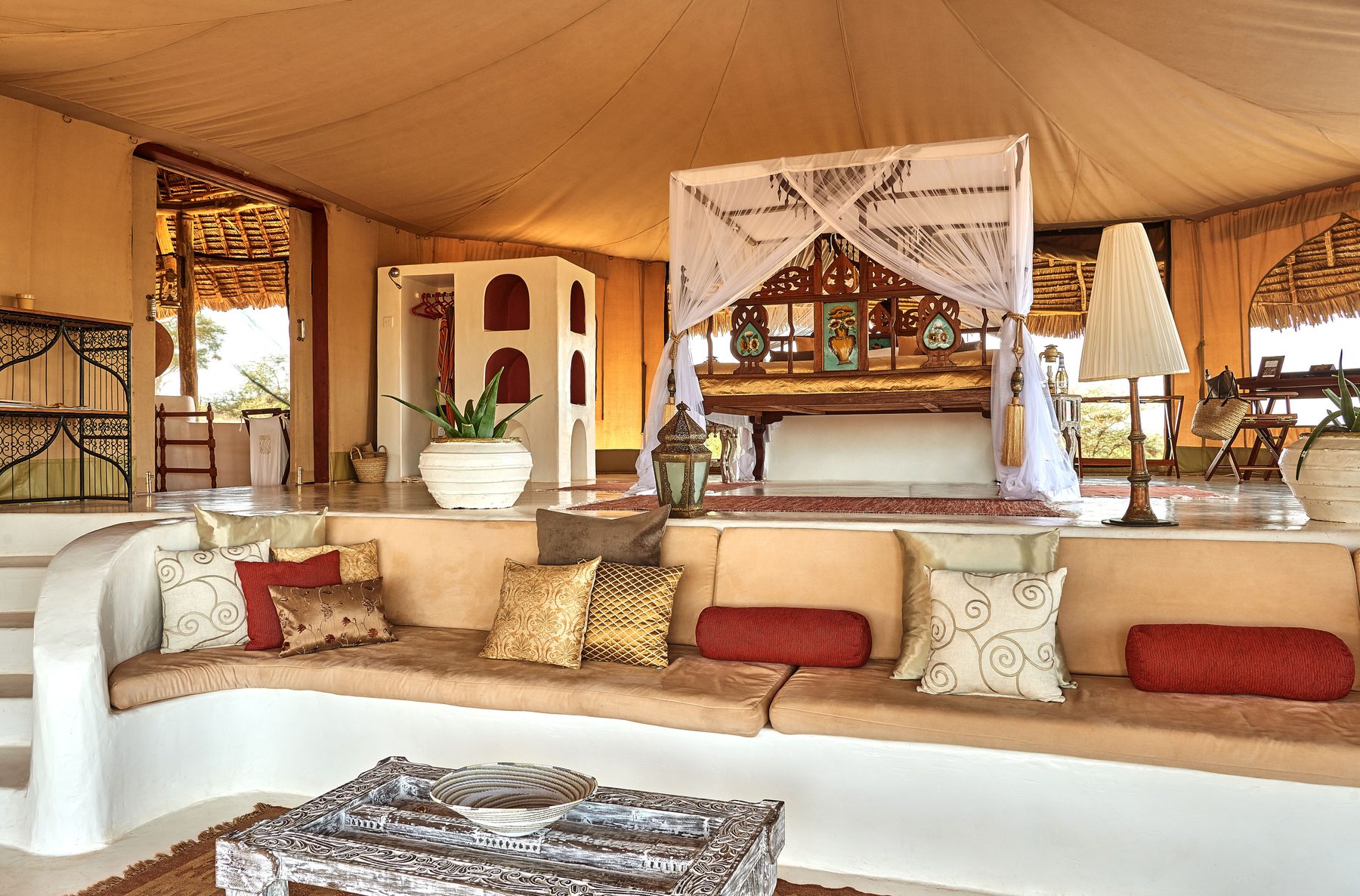 Room at Sasaab with a canopied bed behind a long white built-in sofa scattered with red and gold cushions