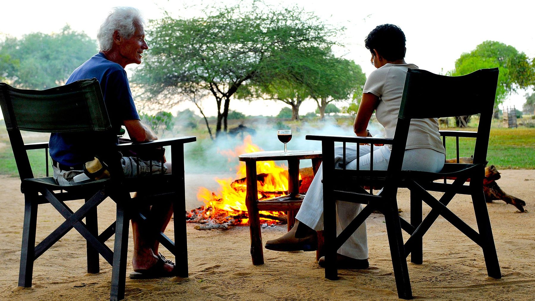 Two guests on canvas chairs with drinks beside a campfire on the sand at Satao Camp