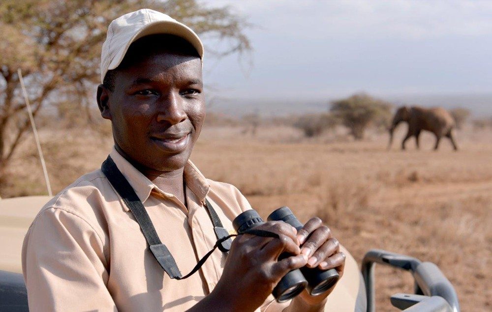 Smiling guide holding binoculars on a game drive from Satao Elerai, with an elephant on the plains behind