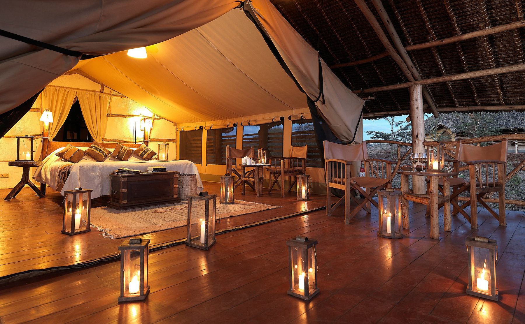 Tent at Satao Elerai lit by candle lanterns, with a double bed inside and safari chairs on a veranda under a thatched roof