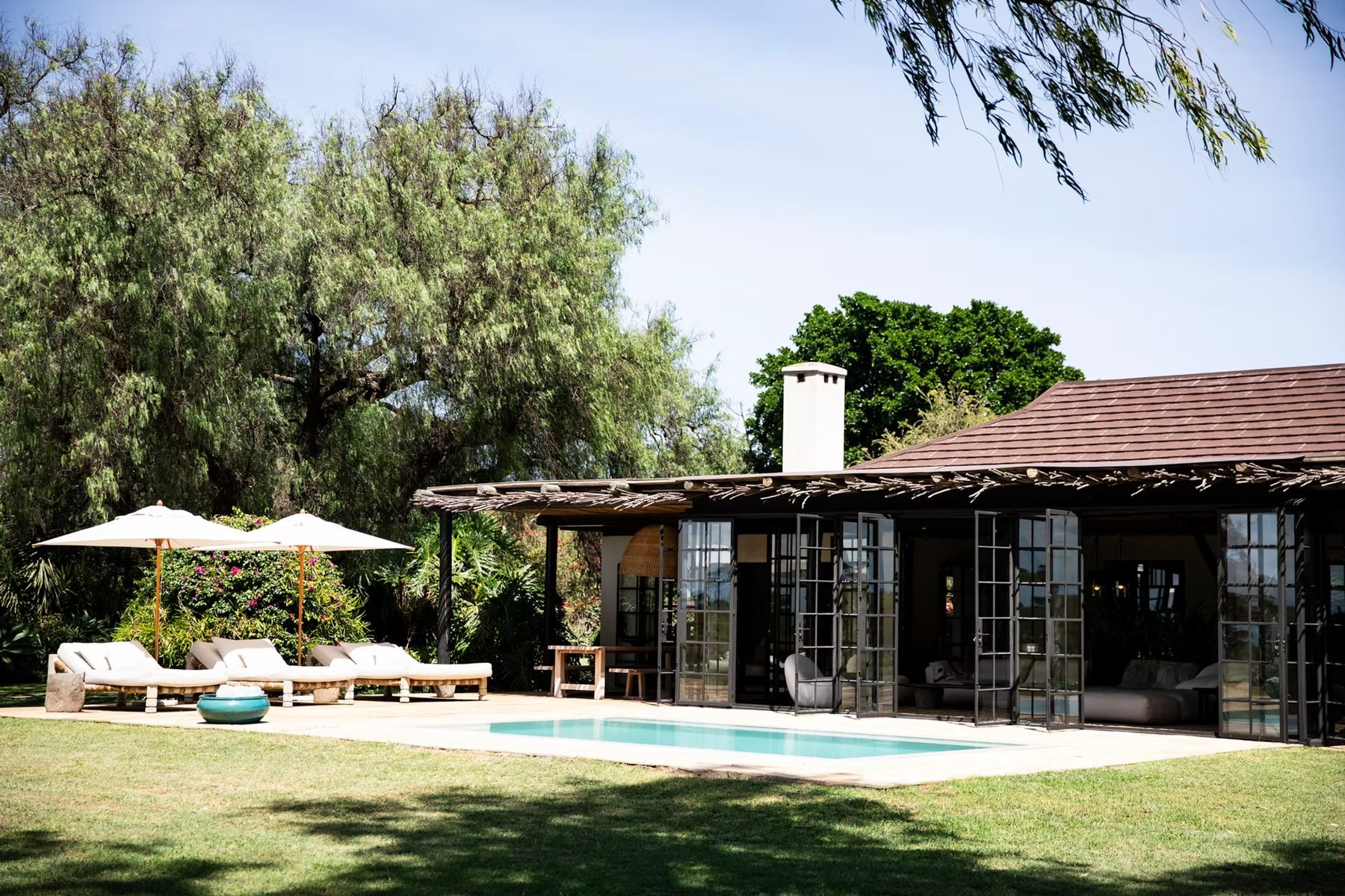 Swimming pool on a wide lawn in front of a glass-fronted house with umbrellas and loungers at Segera Retreat