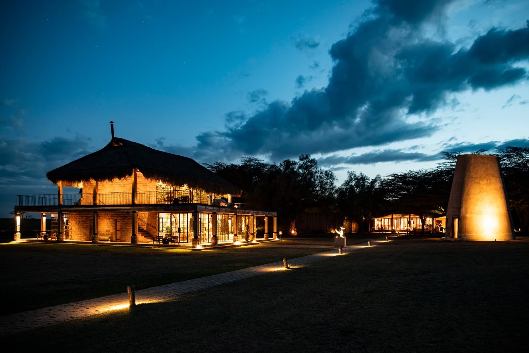 Thatched main house and a lit tower glowing at dusk under a deep blue sky at Segera Retreat