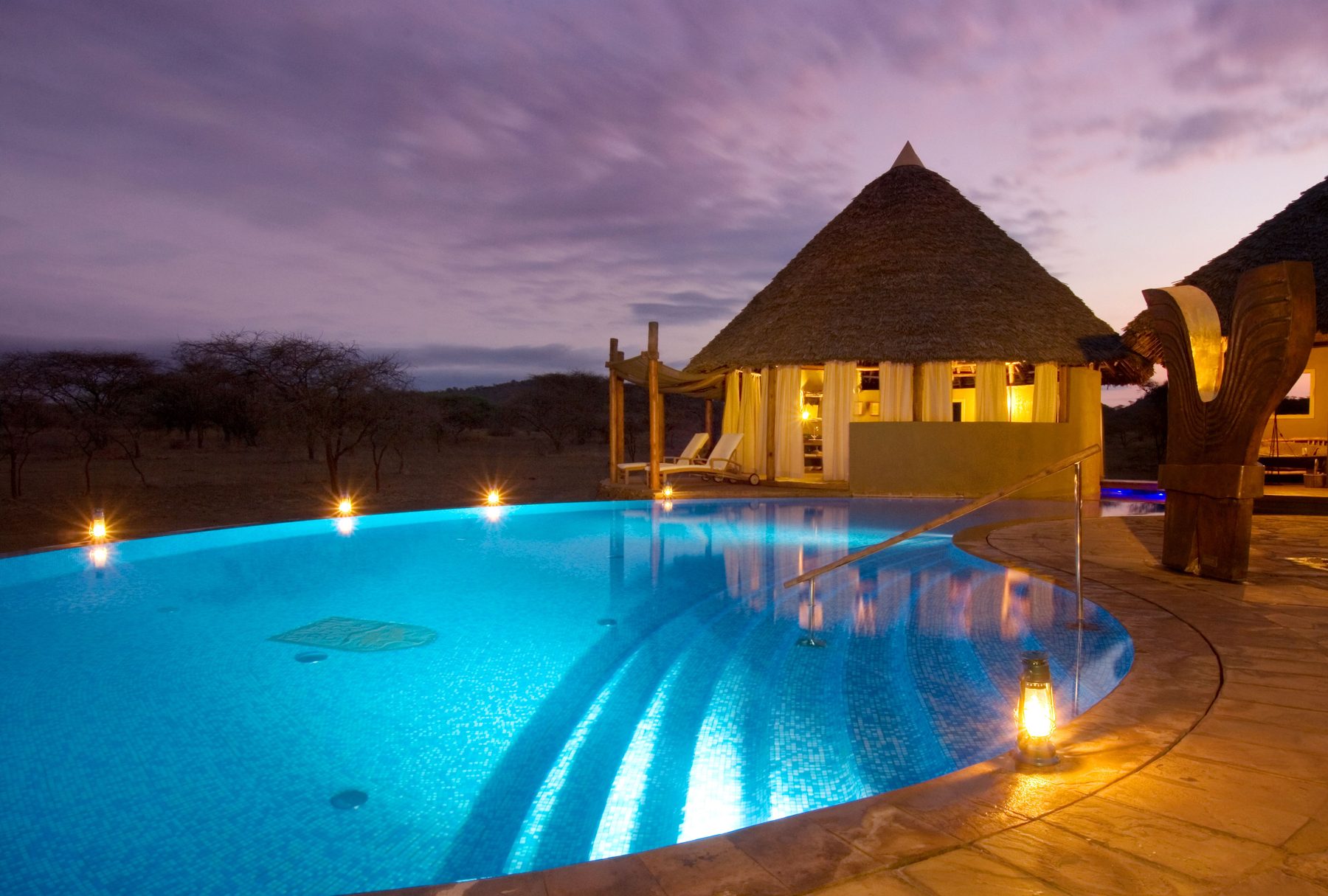 Lit pool beside a thatched building at Severin Safari Camp, with lanterns around the edge at dusk