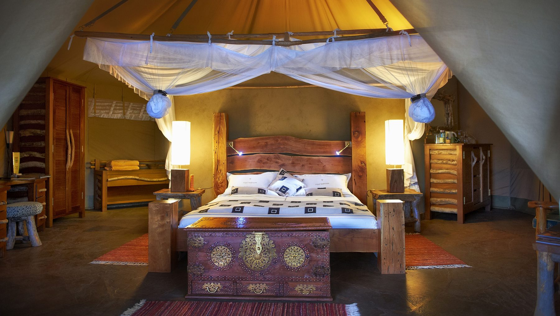 Lamplit tent at Severin Safari Camp with a carved bed, a wooden chest and a high canvas ceiling