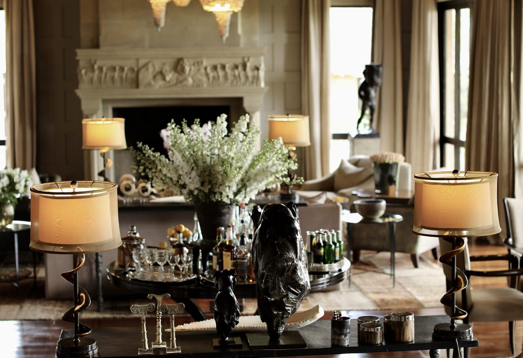 Drawing room at Sirai House with lamps, white flowers and sculptures on low tables before a stone fireplace