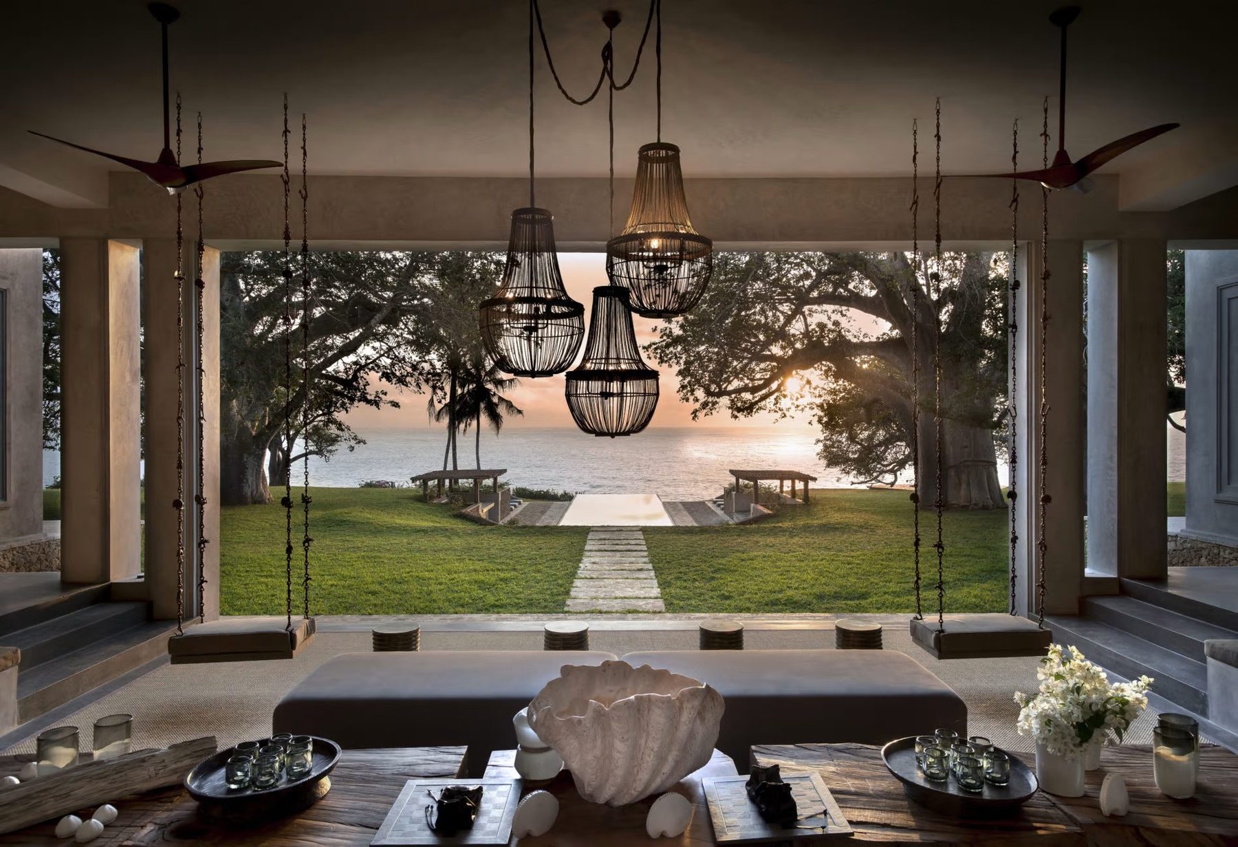 Lounge at Sirai House with chandeliers and low seating, opening onto a lawn and path framed by trees