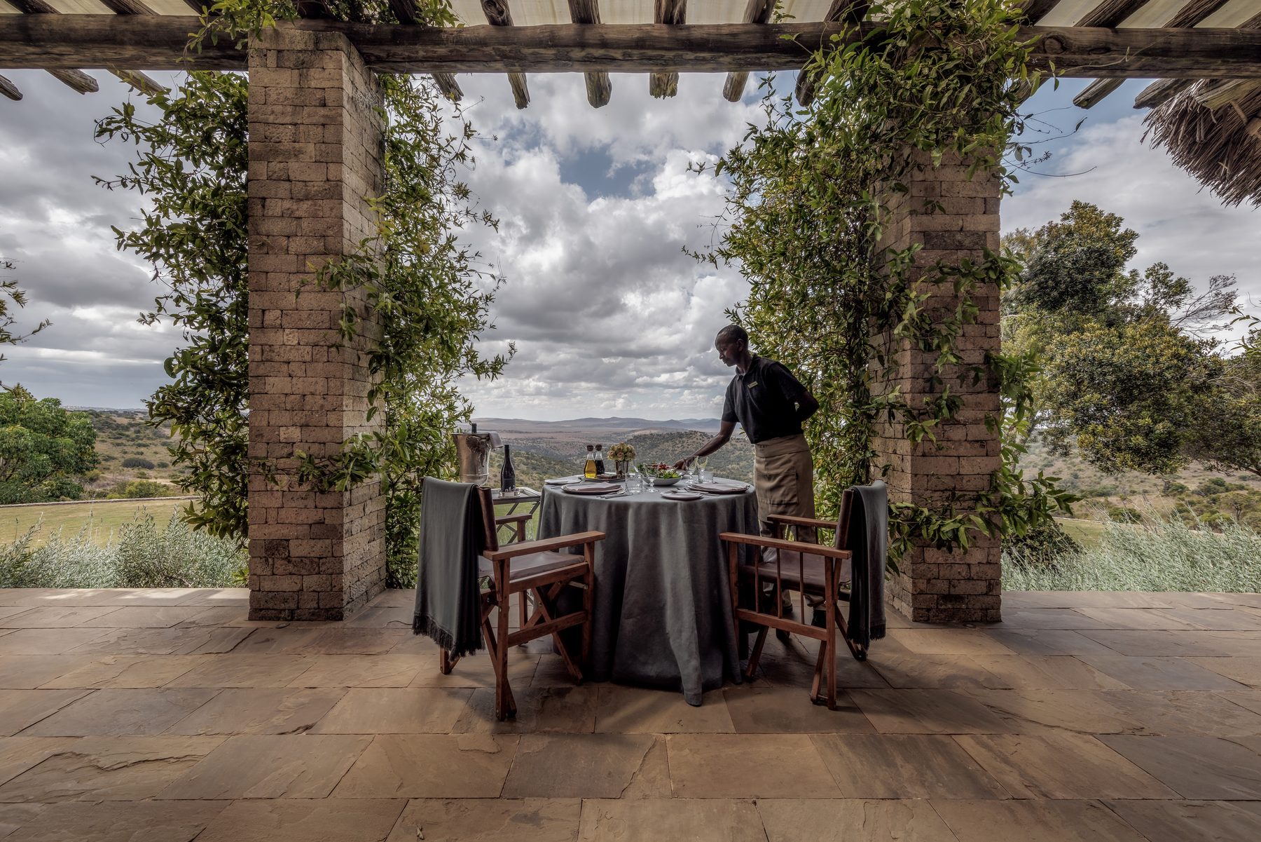 Table being set on a stone terrace under a vine-covered pergola at Sirai House, looking over hills
