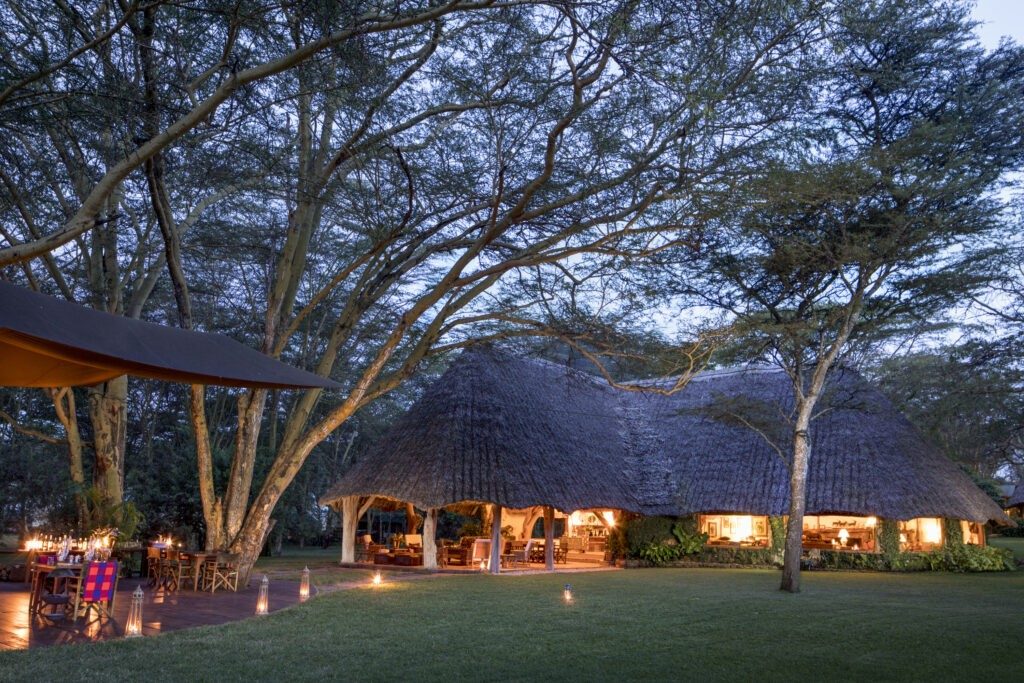 Thatched main house at Sirikoi glowing at dusk beneath tall acacias, with candles on the lawn