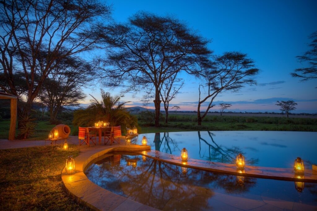 Swimming pool at Sirikoi edged with lanterns at dusk, with acacia trees against a deep blue sky