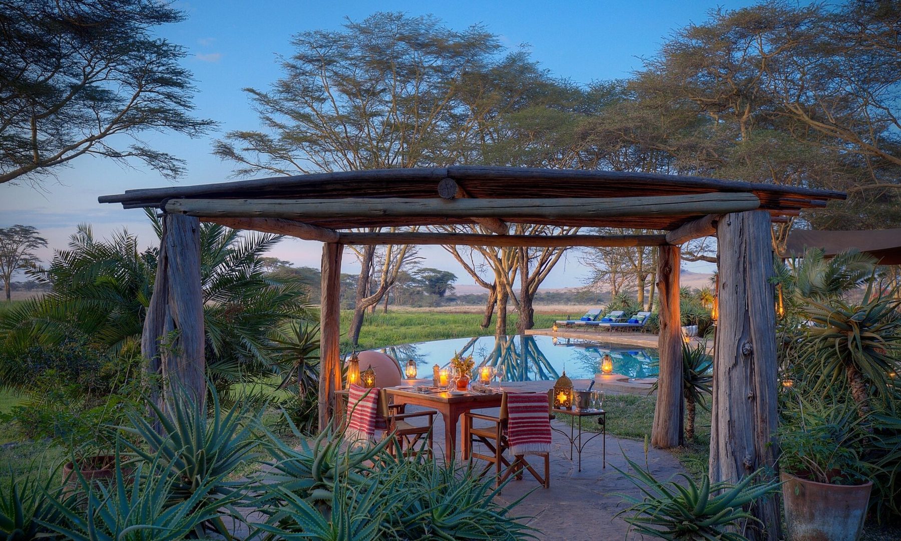 Wooden gazebo with a dining table beside the pool at Sirikoi, surrounded by aloes and acacia trees