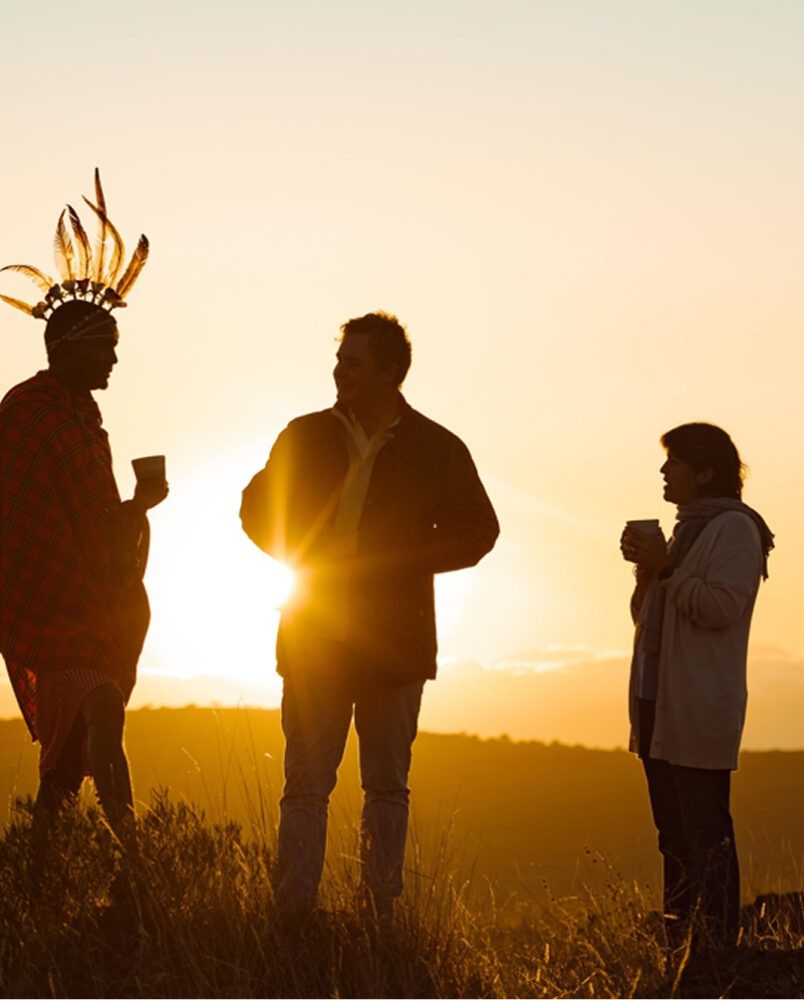Guests silhouetted with drinks at sunset on Lewa, joined by a man in a feathered headdress