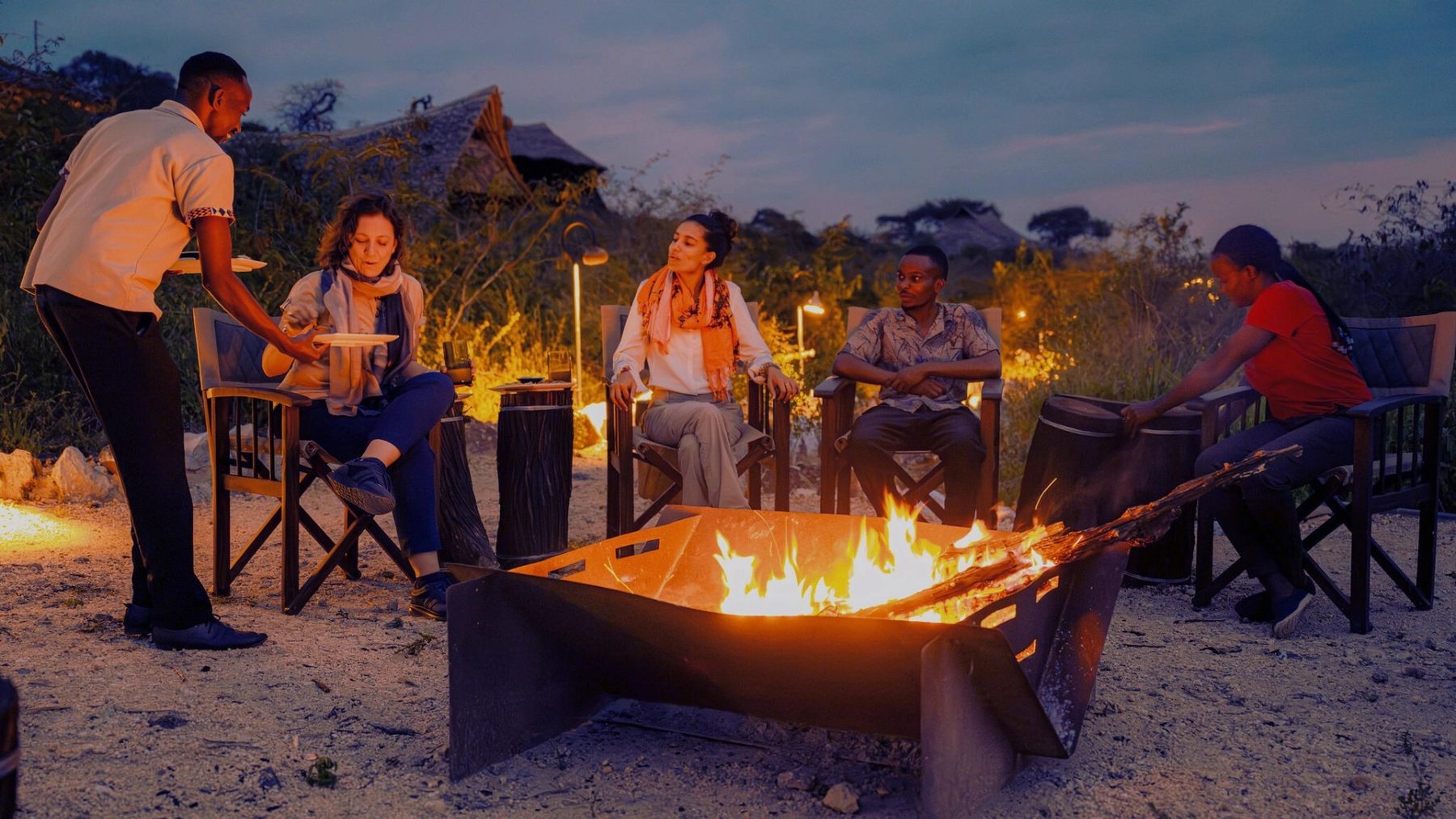 Guests in safari chairs around a fire pit at dusk at Soroi Cheetah Tented Camp, served by staff