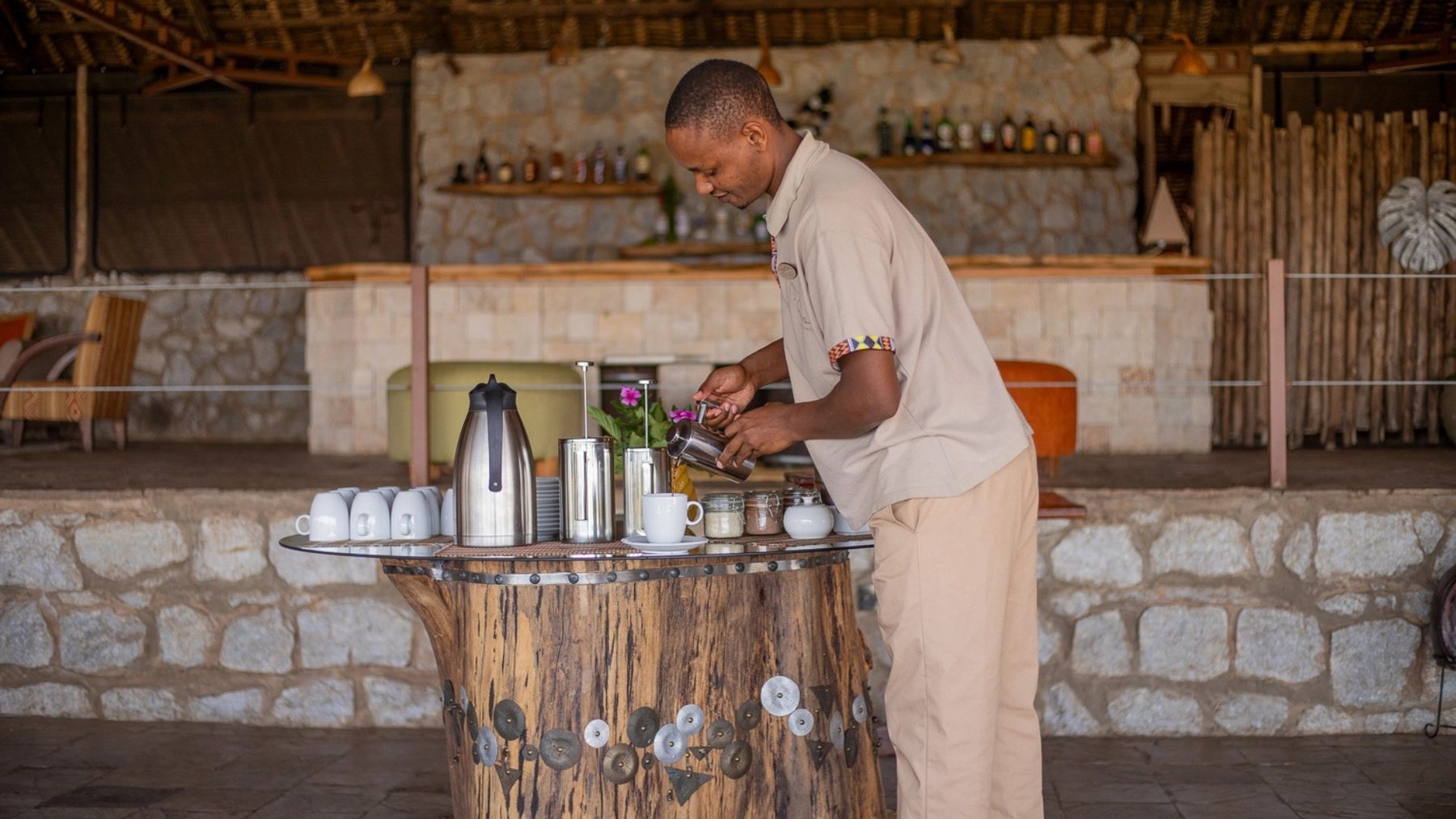 Staff member preparing coffee at a counter in front of the stone-walled bar at Soroi Cheetah Tented Camp