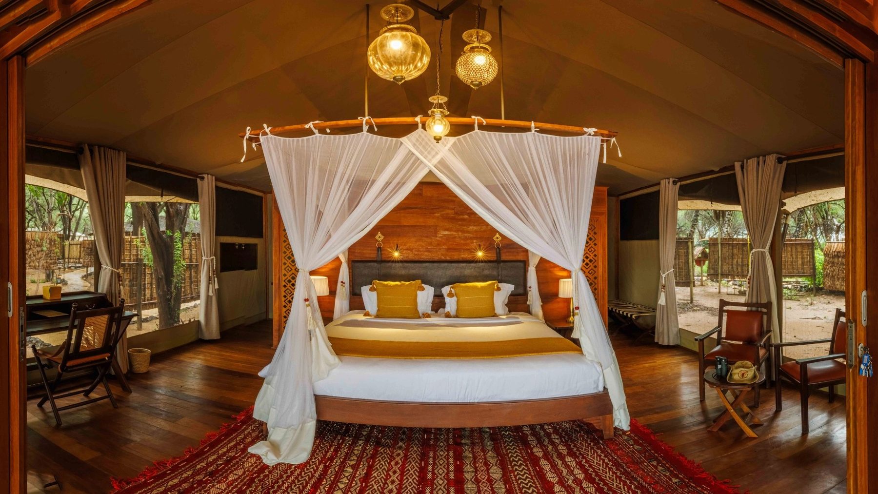 Canopy bed draped in white netting below brass lanterns on a red rug, with leather chairs and a desk, at Soroi Larsens Camp