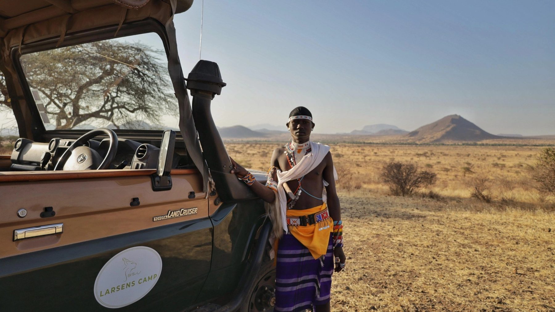 Samburu guide in a beaded collar beside a safari vehicle on dry plains below a pointed hill, on a game drive from Soroi Larsens Camp