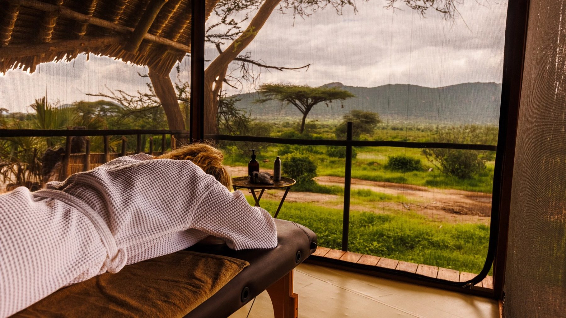 Guest in a robe on a massage bed in a screened room at Soroi Larsens Camp, looking out to acacias and hills