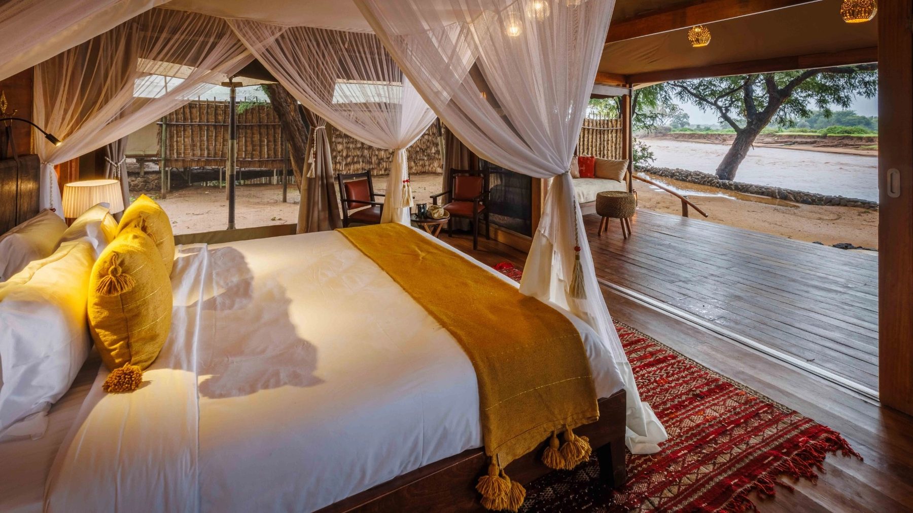 Bed with gold cushions and a netted canopy beside wide doors opening onto a terrace above the river at Soroi Larsens Camp