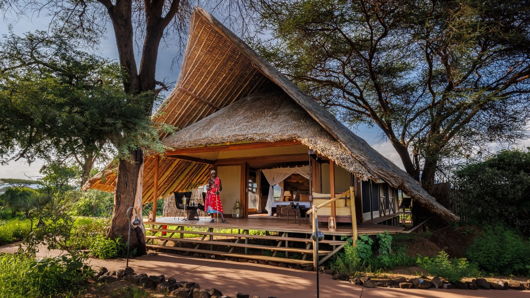 Tent with a tall thatched roof and wooden steps up to its veranda among shady trees at Soroi Larsens Camp