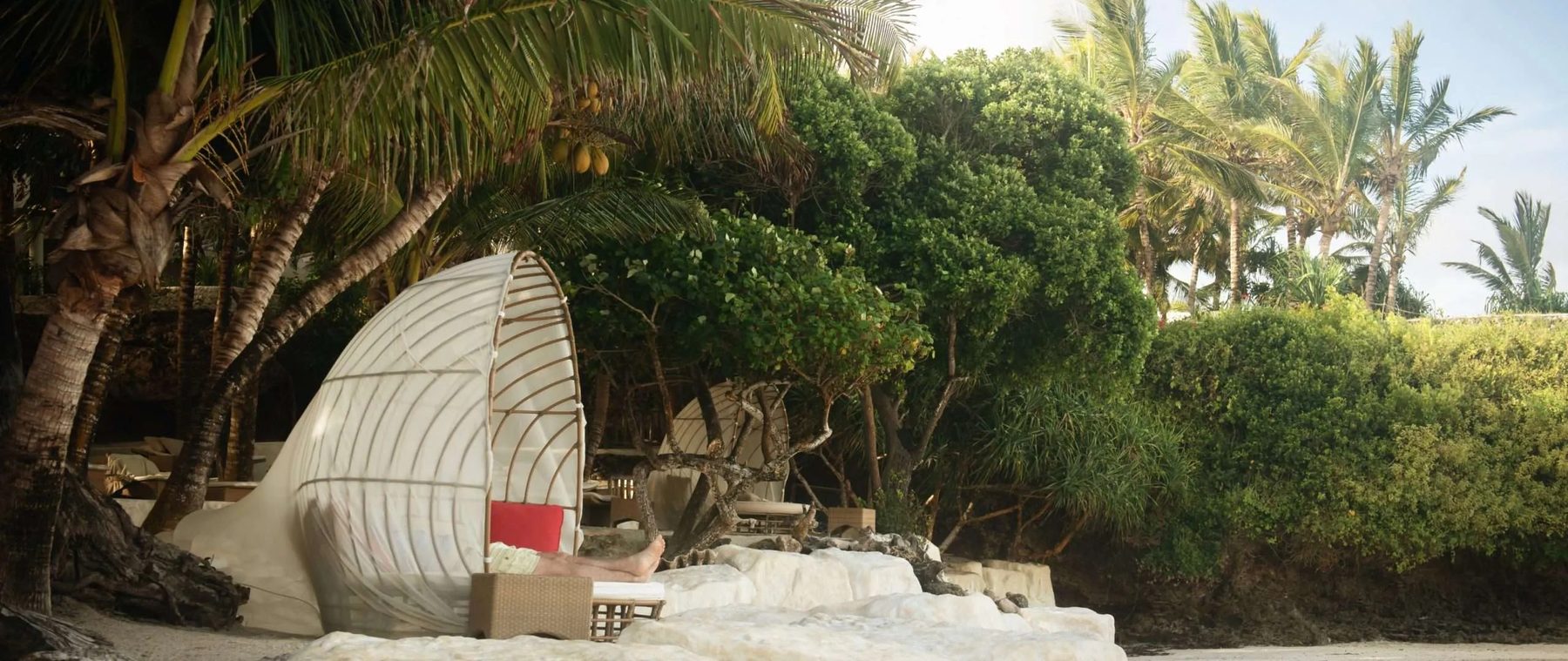 Woven hooded daybed with red cushions on the sand in front of coastal greenery at Swahili Beach Resort