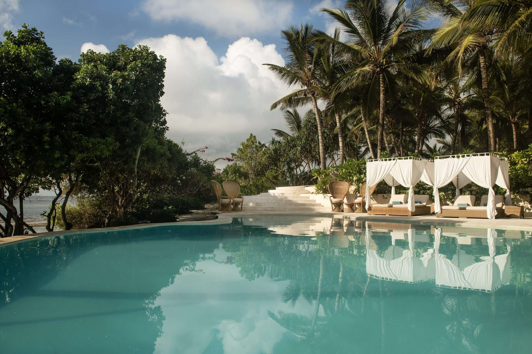 Pool at Swahili Beach Resort reflecting palm trees, with white curtained cabanas along its edge