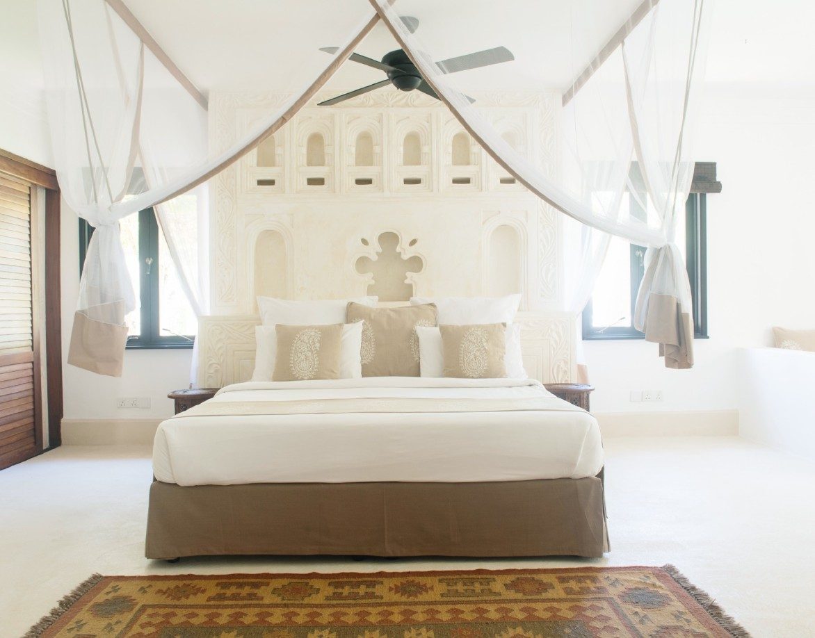 Bright white room at Swahili Beach Resort with a canopy bed, carved plaster headboard wall and a ceiling fan