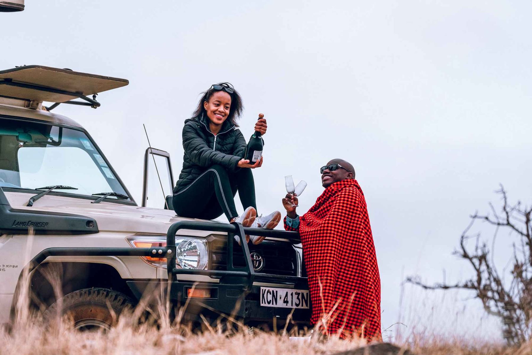 Guest standing in an open game drive vehicle beside a Maasai guide in a red shuka, sharing a drink
