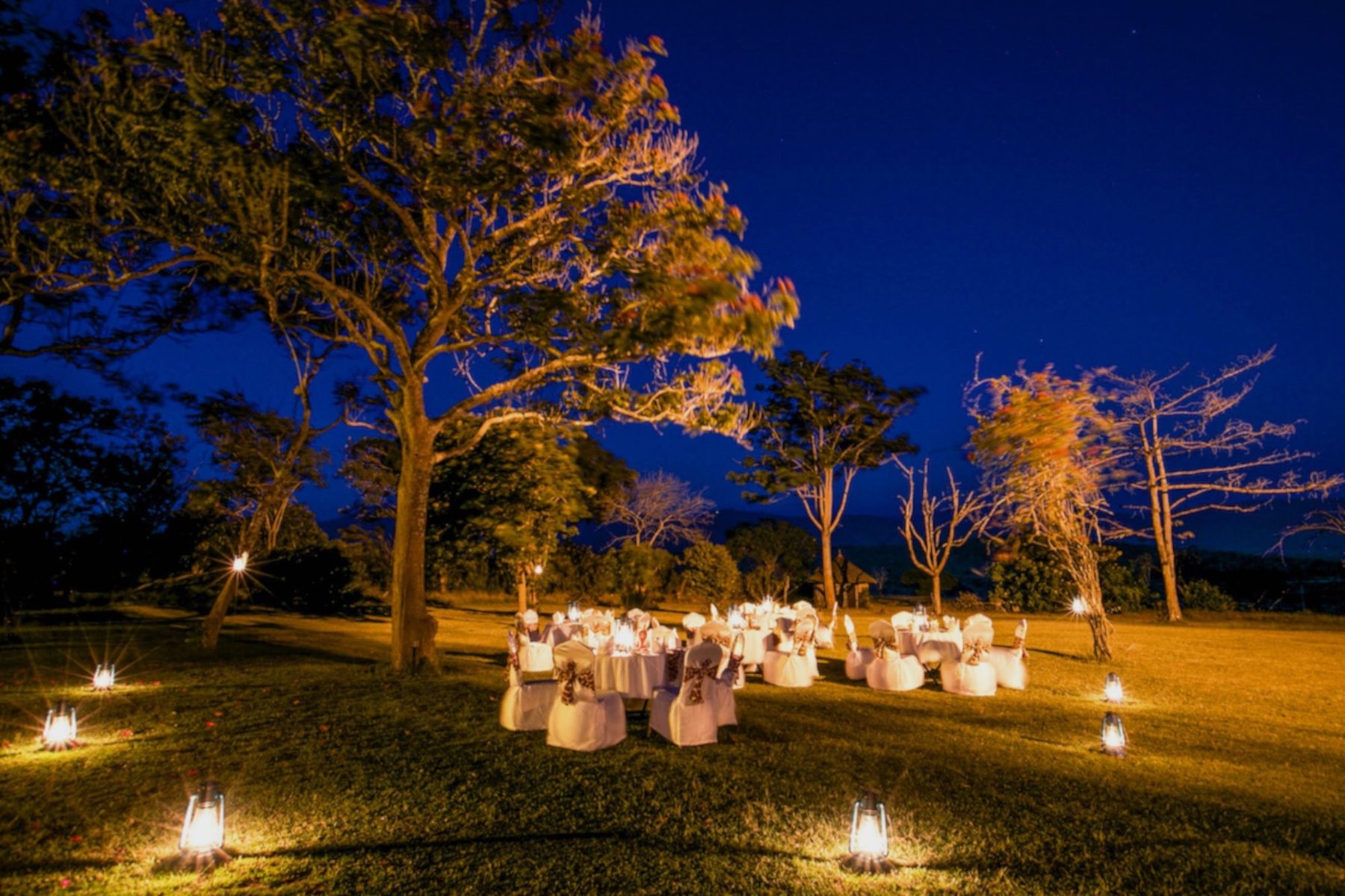 Dinner tables set on a lawn lit by lanterns under trees at dusk at Taita Hills Safari Resort & Spa