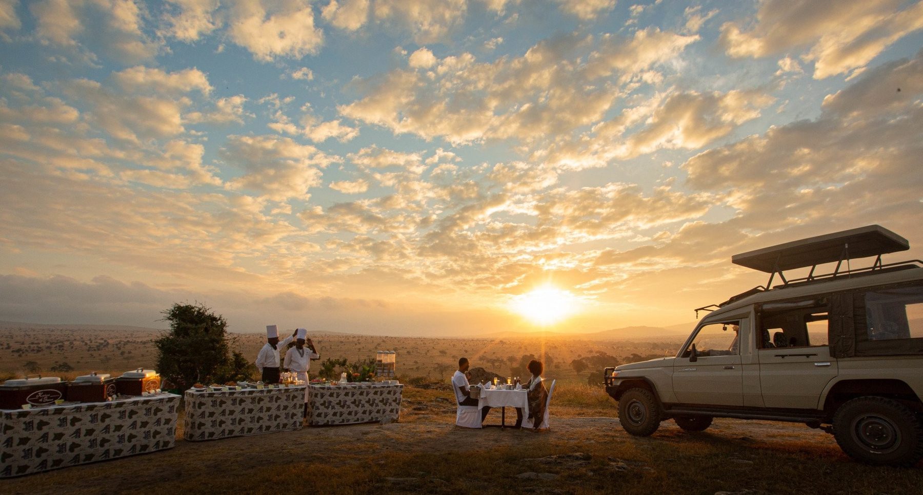 Guests gathered beside a game drive vehicle on a rise, watching the sun set over the plains