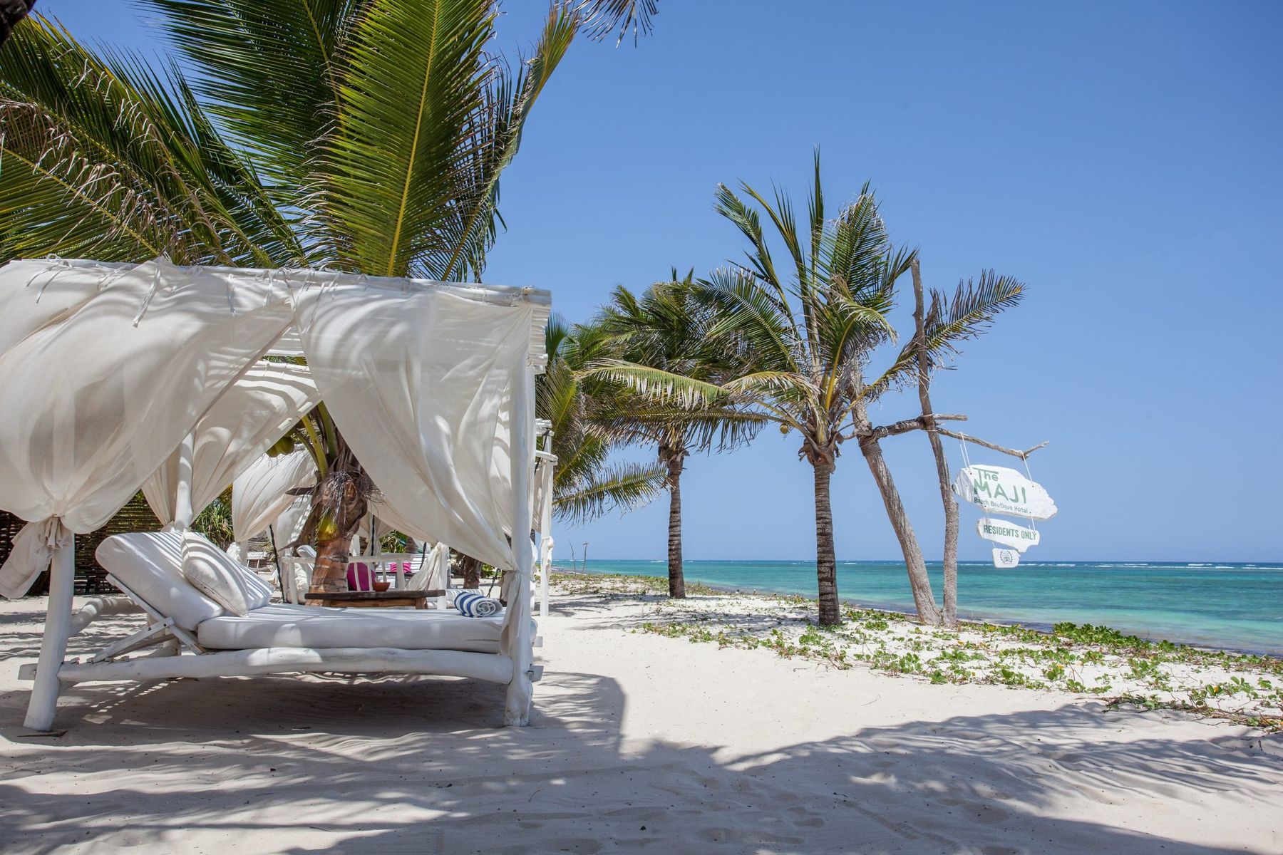 A white canopied day bed under palm trees on the sand at The Maji, with the turquoise sea beyond