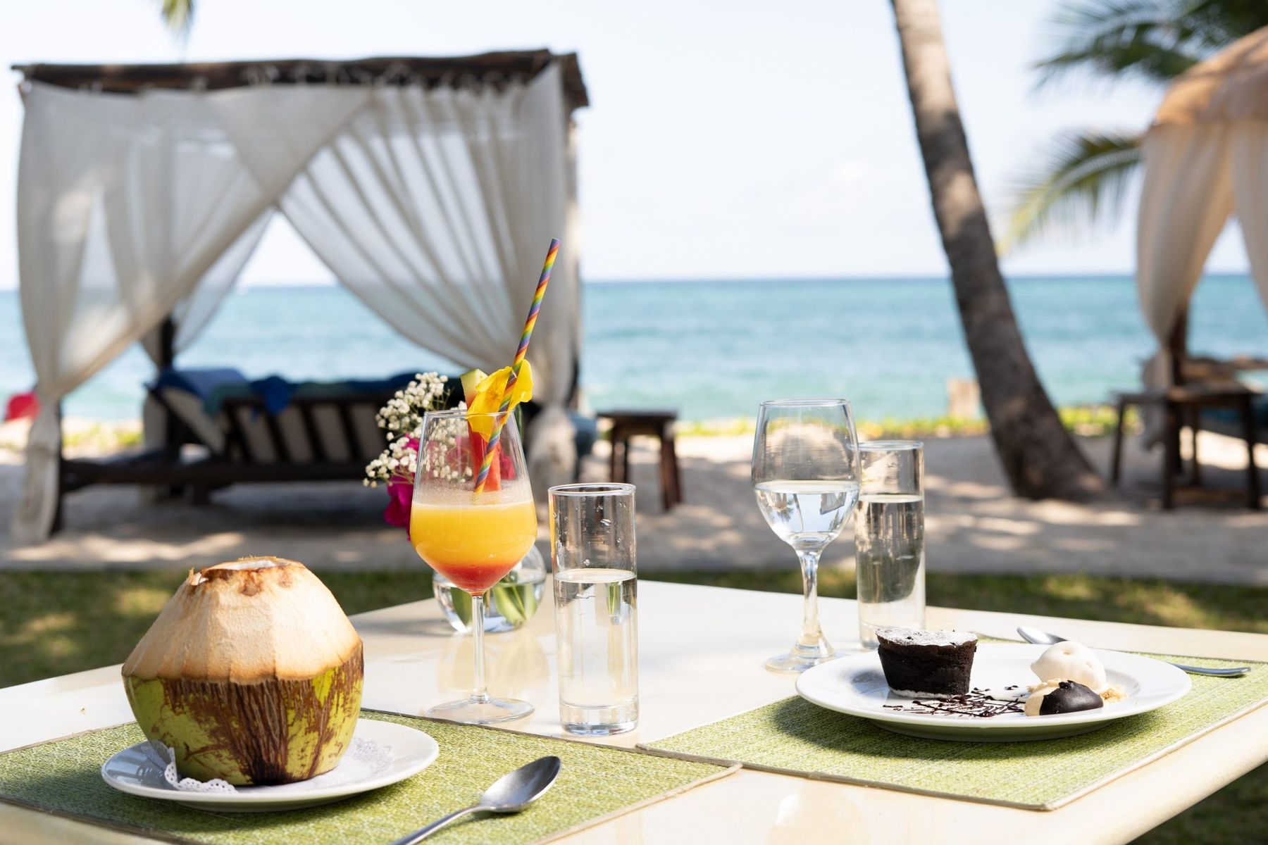 A fresh coconut, a cocktail and dessert on a table by the beach at The Maji