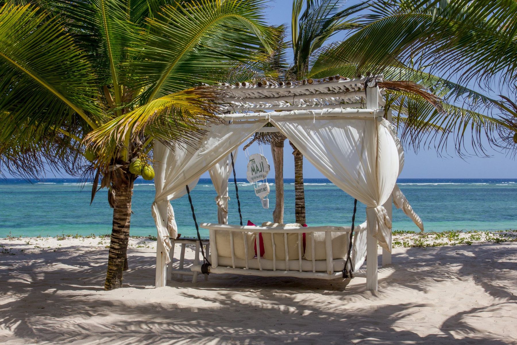 A cushioned swing seat under a white wooden canopy on the beach at The Maji, between palms and the sea