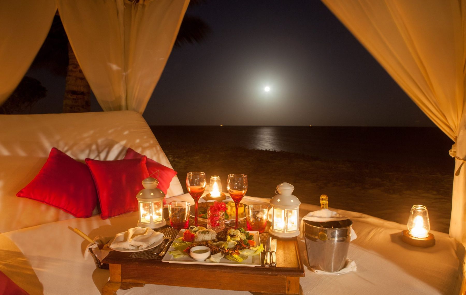 Candles, cushions and a tray of food in a curtained cabana facing the moonlit sea at The Maji