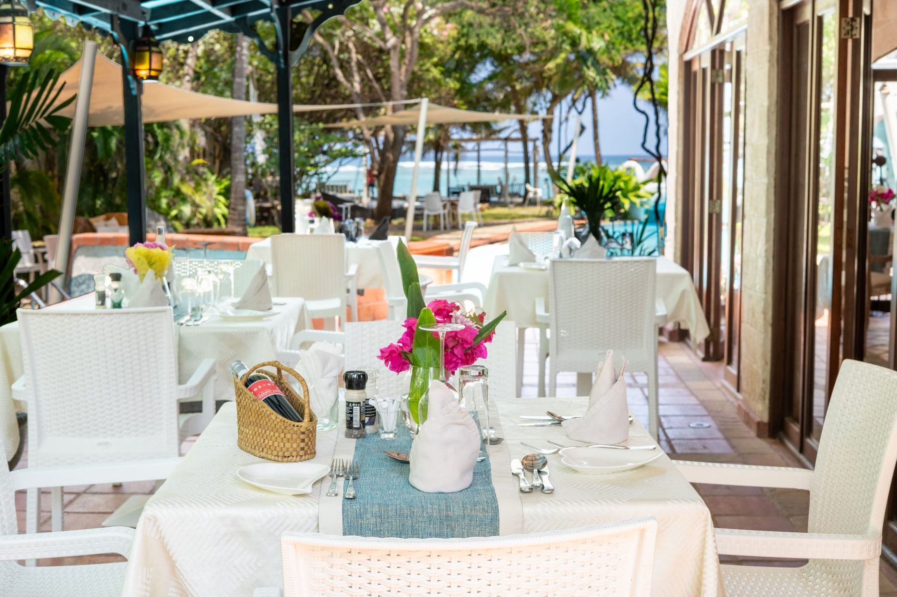 Tables laid with white cloths and flowers on the shaded restaurant terrace at The Maji, with the beach beyond