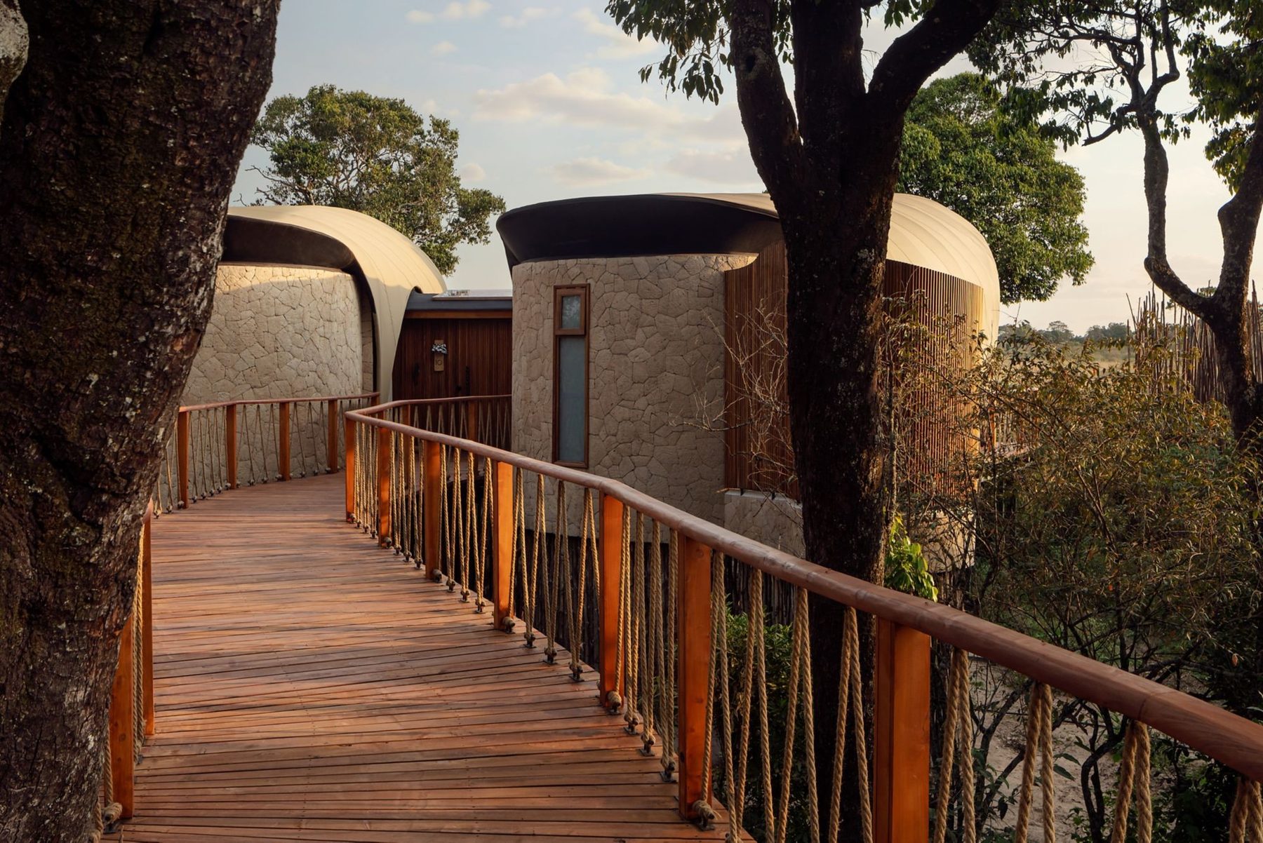 Raised wooden boardwalk leading between round stone-walled buildings among trees at The Ritz-Carlton, Masai Mara Safari Camp