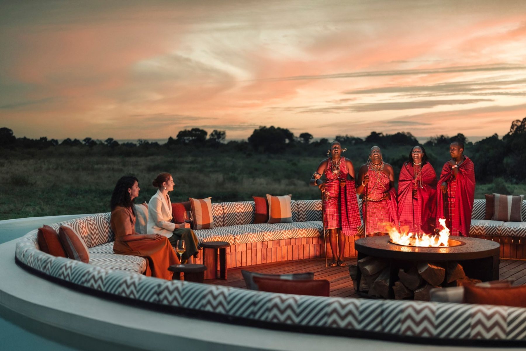 Guests seated around a fire pit at dusk at The Ritz-Carlton, Masai Mara Safari Camp, with Maasai singers in red shukas